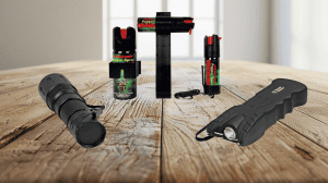 Pepper spray for home, car and personal, a master blaster stun gun and a tactical flashlight all displayed on a wooden table with light from a window in the rear of the image.